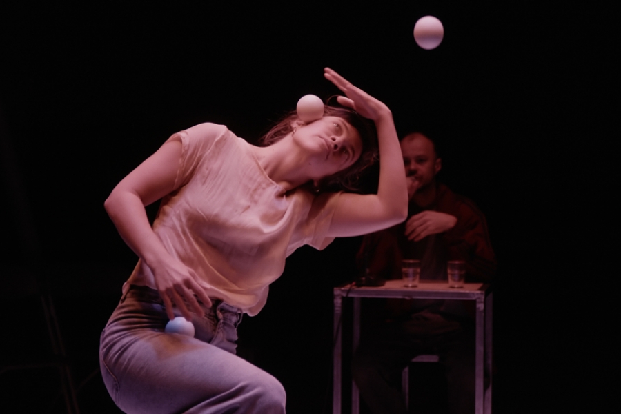 Suc scène, la jongleuse a la tête penchée et fait tenir en équilibre une balle. La deuxième balle est dans les airs et la troisième dans sa main. à l'arrière plan, en floue, il y a le rappeur à une table.