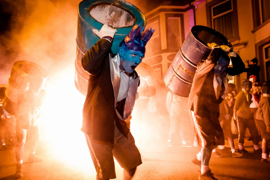 Dans la rue, les interprètes déambulent. Au premier plan on voit deux interprètes, le visage peint en bleu avec des coupes de cheveux excentriques. ils portent des tonneaux sur le dos. Derrière eux, un feu ou une explosion.