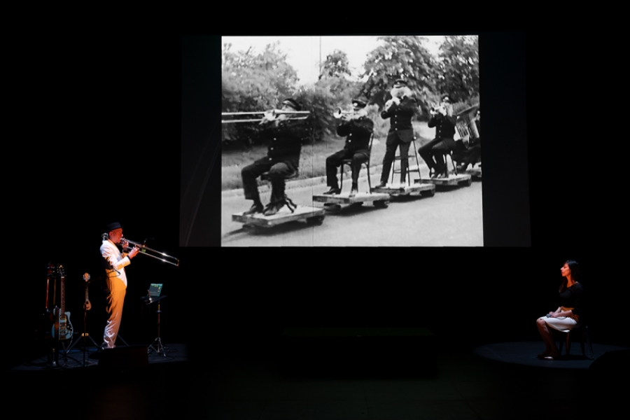 Un musicien joue du trombone sur scène, plusieurs instruments sont disposés à côté de lui. Une grande projection en noir et blanc est visible en fond, représentant une fanfare.