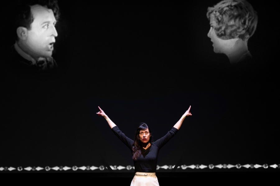 Une comédienne est debout, bras levés, au centre de la scène, avec une projection derrière elle montrant deux visages issus d’un film muet.