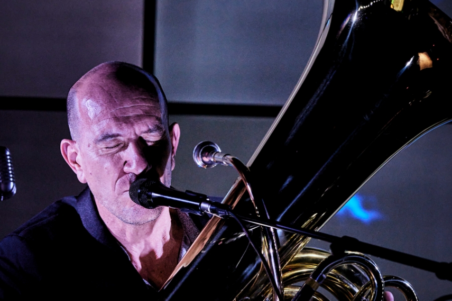 Le même homme joue du tuba, concentré, les yeux clos, les lèvres collées au micro comme s’il murmurait une note grave. Les reflets métalliques de l’instrument captent la lumière, amplifiant l’impression de puissance contenue.
