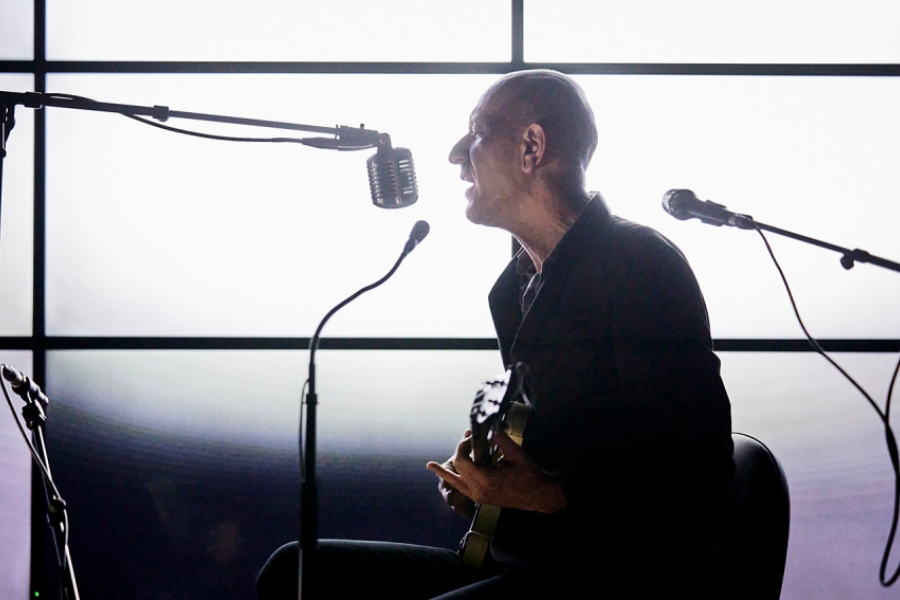 Silhouette sombre en contre-jour, l’homme est assis, guitare sur les genoux, chantant dans un micro suspendu comme dans une vieille émission de radio. L’arrière-plan blanc et lumineux découpe sa posture tendue et expressive.