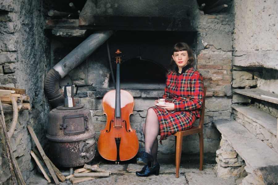 L'artiste est assise sur une chaise en bois dans le salon d'une vieille maison de campagne abandonnée. Elle est en robe rouge et à côté d'elle se trouve son violoncelle. 