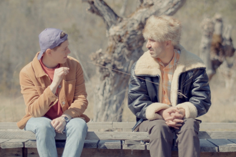 En pleine nature, sur un banc en bois, deux hommes sont assis et se parlent. Celui de gauche porte une casquette mauve. Celui de droite porte une chapka à fourrure et un manteau en cuir.