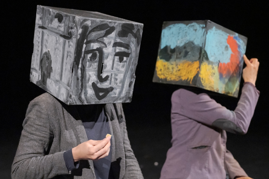 Deux interprètes sont assis, chacun avec une boîte cubique peinte sur la tête. Celle de gauche est monochrome, représentant un visage stylisé en noir et blanc, tandis que celle de droite est couverte d’un motif abstrait coloré (bleu, jaune, rouge). Le contraste entre les deux boîtes crée un jeu visuel saisissant, entre figuration et abstraction. Les costumes sobres mettent en valeur l’aspect graphique des têtes cubiques.
