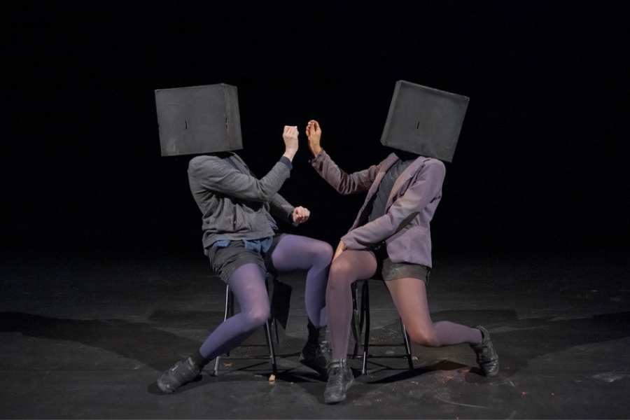 Les deux performeurs, toujours coiffés de boîtes noires anonymes, sont assis face à face sur des chaises, mimant une gestuelle théâtrale qui évoque un dialogue ou une confrontation silencieuse. Leurs bras tendus, presque synchronisés, dessinent une symétrie dynamique. La scène, épurée et plongée dans l’obscurité, accentue la dimension chorégraphique et énigmatique de l’échange.