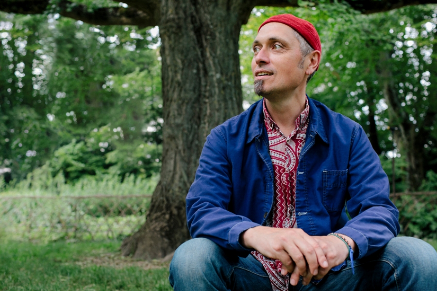 Dans la nature verdoyante, Thierry Combe est assis, au premier plan. Il porte une chemise bleu et un bonnet rouge. Il tourne la tête en rendant vers sa droite.