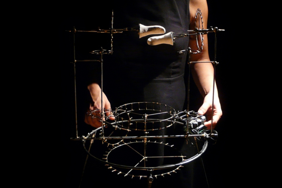 Plan serré sur une structure mécanique à rouages, tenue entre les mains de l'interprète. L'objet semble tourner lorsqu'on l'actionne.