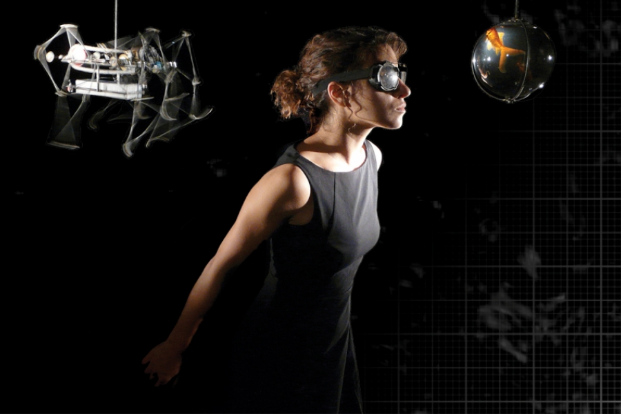 L'interprète porte des lunettes de protection - aviation, elle est tournée de trois quart en direction d'une boule mécanique suspendue, à gauche. À droite, une construction mécanique est suspendue aussi.