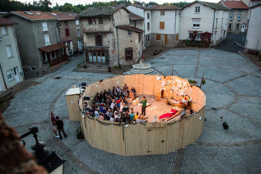Vue aérienne du spectacle se situant sur la place d'un village. On voit bien la scénographie circulaire et en bois, avec le public à l'intérieur.