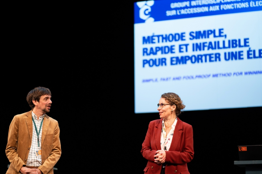 Les deux mêmes intervenants, debout face au public, échangent avec le sourire devant un écran affichant un titre en lettres majuscules : "Méthode simple, rapide et infaillible pour emporter une élection". L’ambiance est à la fois sérieuse et légèrement ironique, évoquant une conférence théâtralisée.