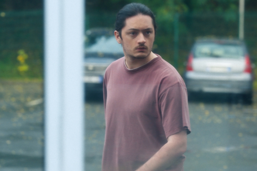 À travers une fenêtre vitrée, on voit un homme en t-shirt rouge qui est dehors, qui cherche à regarder à l'intérieur. 
