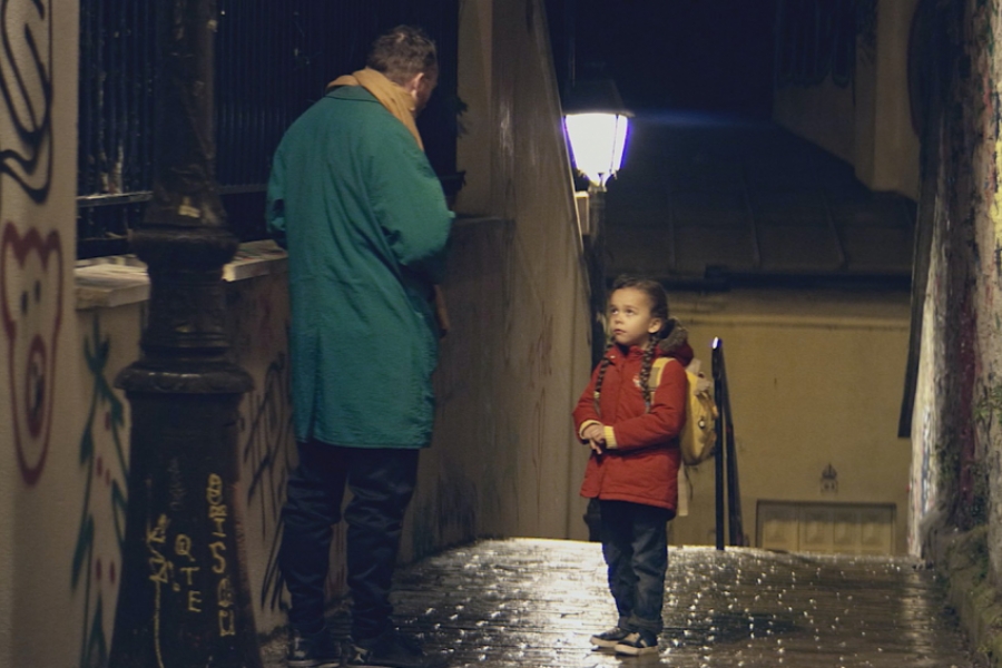 Dans une ruelle sombres, une enfant avec son sac à dos est face à un homme en manteau vert. On ne voit pas le visage de l'homme.