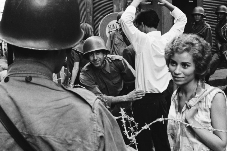 Photo en noir et blanc. Une femme tournée de trois quart et derrière des barbelés, regarde un soldat, qui est dos à la caméra.