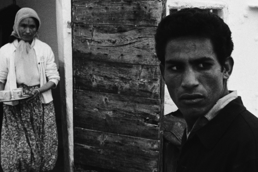 Photo en noir et blanc. Au premier plan à droite, un homme regarde sur la droite d'un air inquiet. À l'arrière plan, une femme marche en portant un plateau avec deux boissons.