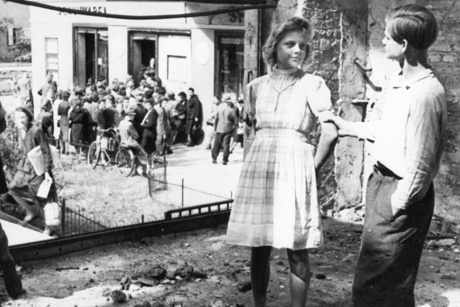 Photo en noir et blanc. Au premier plan, un garçon et une fille parlent entre eux dans des ruines. À l'arrière plan, un regroupement d'adultes devant un bâtiment.