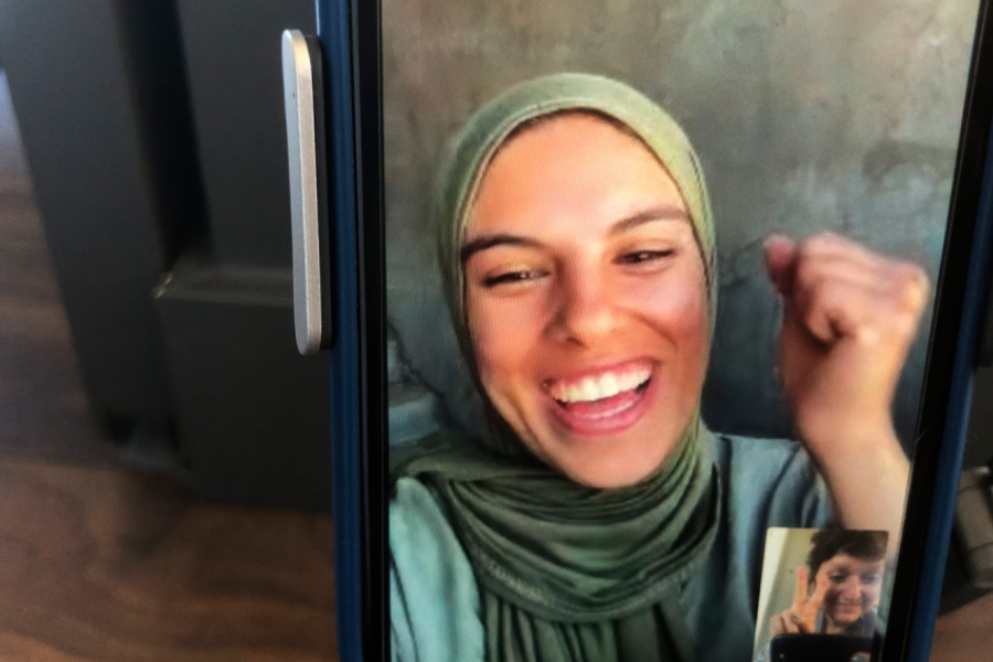 Un smartphone est posé sur un table, on voit la jeune photojournaliste Fatma Hassona en FaceTime avec Sepideh Farsi. Fatma sourit et lève le poing, déterminée. 