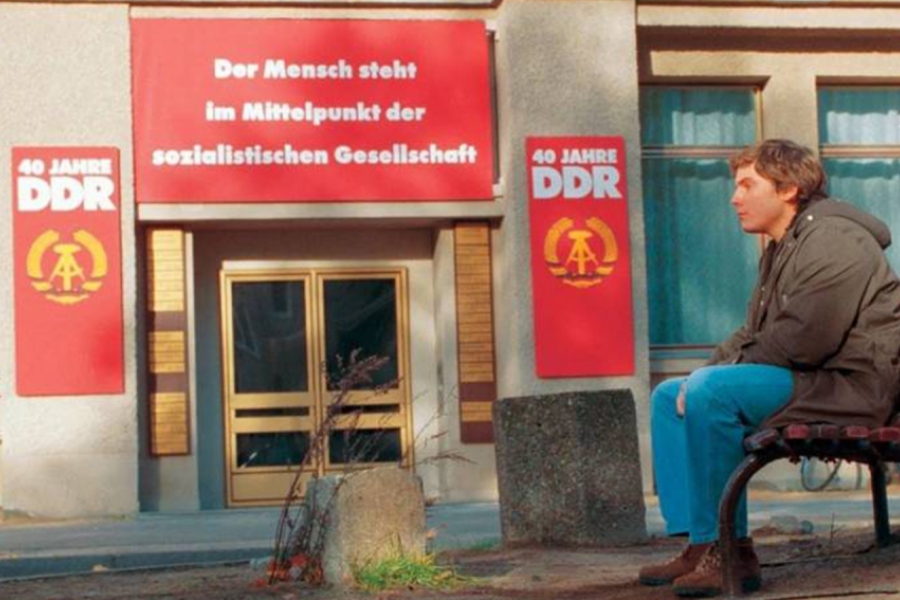 Le fils est assis dehors sur un banc, face à un ancien magasin de la DDR (Deutsche Demokratische Republik).