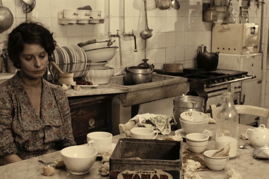 La femme est assise dans sa cuisine. Face à elle, la table est remplie de vaisselles. Elle regarde sa tasse en face d'elle, l'air triste.