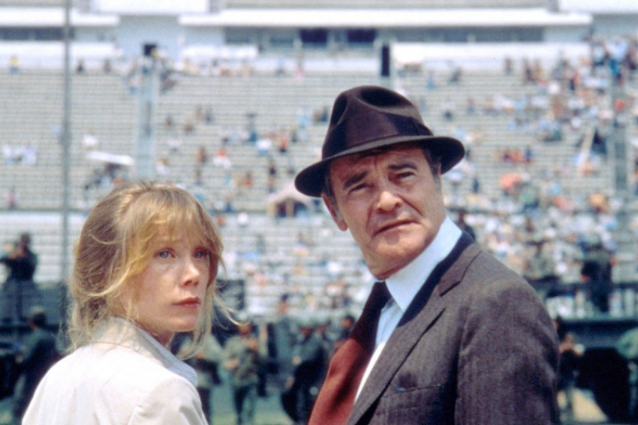 Dans un stade, un homme avec un chapeau et une femme blonde sont tournés de trois quart et regardent derrière eux.