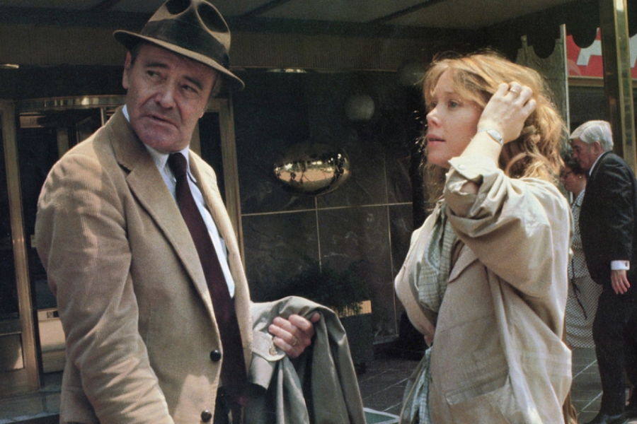 Un homme en costume et avec un chapeau est debout avec une femme blonde devant les portes d'un bâtiment. ils regardent derrière eux.