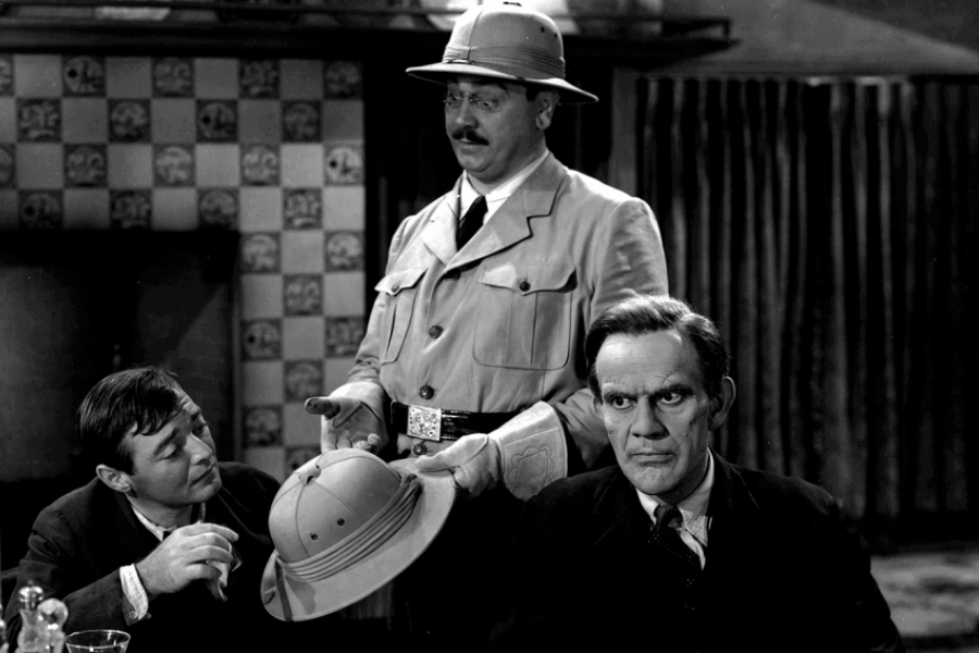 Photo en noir et blanc. Un homme habillé en explorateur se tient debout entre deux hommes assis. Celui de droite est Frankenstein. Celui de gauche à l'air perdu.