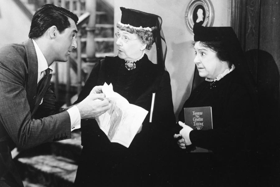 Photo en noir et blanc. Un homme à gauche tend un papier à deux dames vêtues de noir.