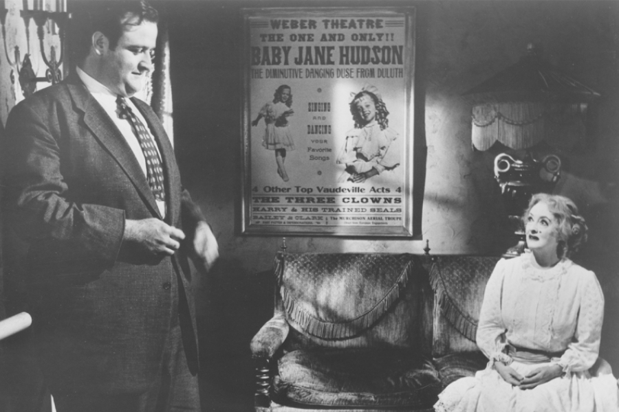 Noir et blanc. Baby Jane est assise sur un canapé et regarde l'homme d'affaire qui est à gauche. Entre eux, une affiche du show de Baby Jane est accroché au mur.