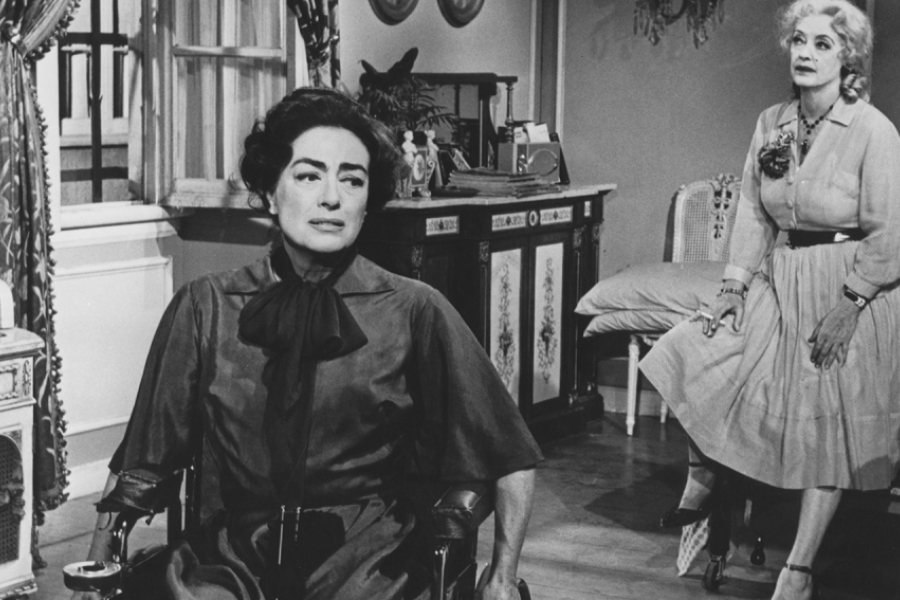 Noir et blanc. Au premier plan, Blanche est assise sur son fauteuil roulant et semble déboussolée. Derrière elle, Baby Jane fume une cigarette assises sur un lit, l'air pensante. 