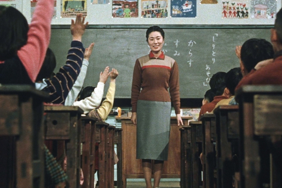 Dans une classe d'école, l'institutrice est placée devant son bureau. Les élèves de la rangée de gauche lèvent tous la main. La rangé des élèves à droite sont tous habillés et coiffés de la même manière.