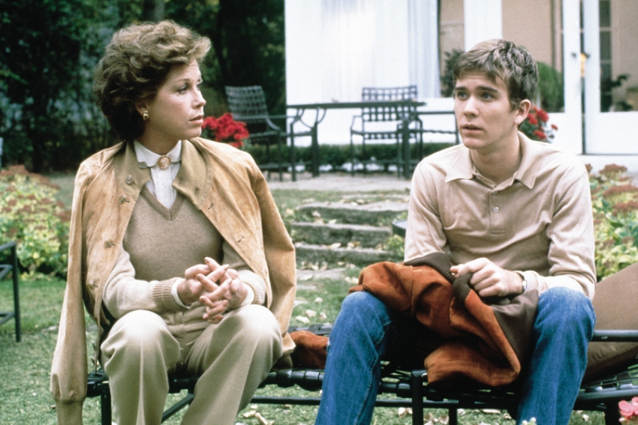 Le jeune garçon et sa mère sont assis sur un banc dehors. Elle le regarde tandis que lui regarde ailleurs.