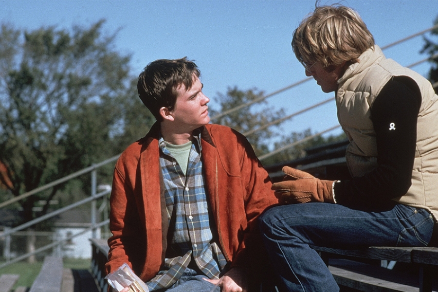 Dans des gradins en extérieur, le jeune garçon parle à un homme qui est de dos.