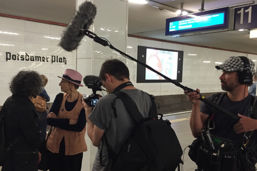 Dans une station de métro, une équipe de tournage suivent des gens, on voit la perche à micro et la caméra.