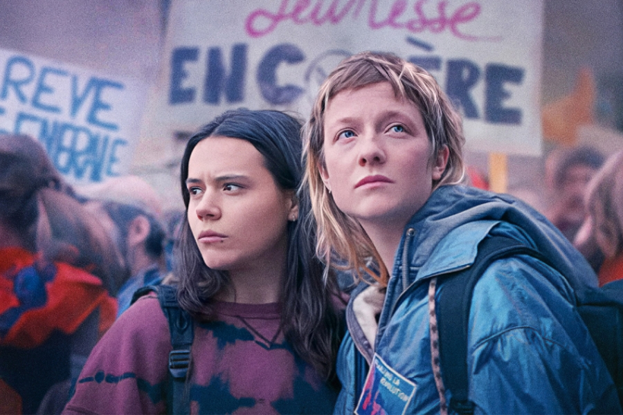 Deux jeunes femmes, une blonde et une brune, sont au centre d'une manifestation étudiante dans la rue. Au fond, on voit des pancartes dans la foule.