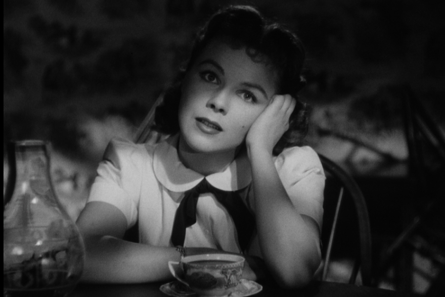 Une femme attablée et la main sur son visage, regarde en face d'elle, l'air songeuse.