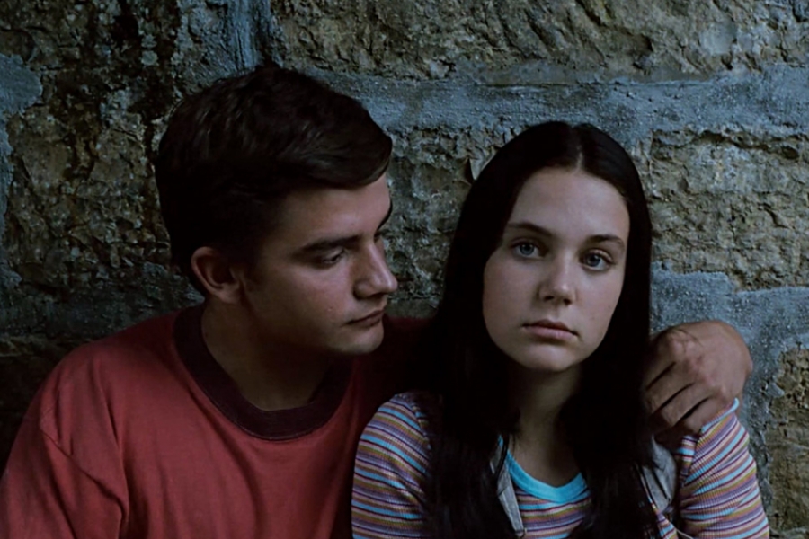 Contre un mur en pierre, un garçon et une fille sont côte à côte. Le garçon regarde la fille. La fille regarde l'objectif