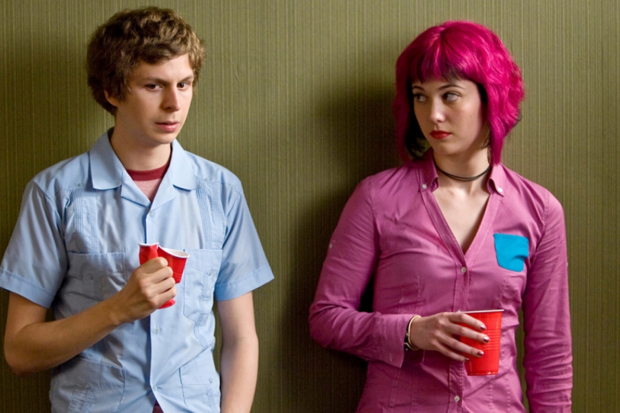 Un étudiant en chemise bleu et à côté d'une étudiante qui est tout en rose (chemise et cheveux). Elle le regarde.