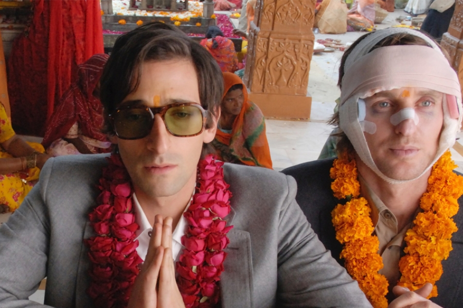 Gros plan sur les deux frères. Ils portent des colliers de fleurs. Celui de gauche porte des lunettes de soleil et un costume gris, il ferme les yeux. Le second à droite porte aussi un costume, mais il a beaucoup de bandages et pansements au visage et autour du crâne. Il a les yeux ouverts et regarde devant lui.