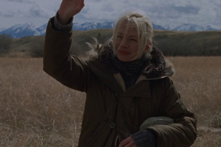 Au premier plan, une femme blonde avec un manteau lève la main en s'adressant à quelqu'un. Derrière elle on voit qu'elle est près d'un champ, au loin il y a des montagnes.