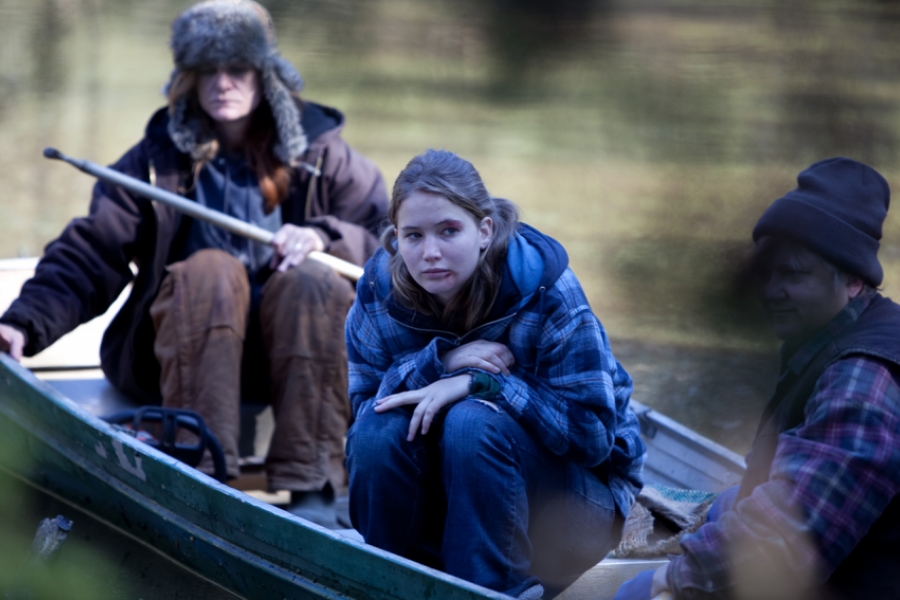 Sur une barque sur un lac, une jeune femme est recroquevillée, entourée de deux hommes qui mènent la barque près du rivage.