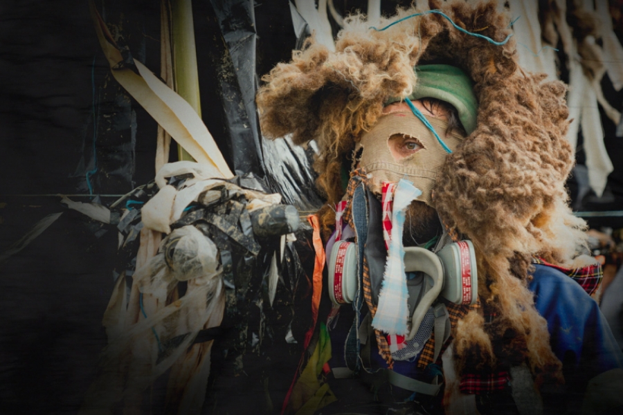Portrait rapproché d’une personne portant un masque improvisé et un filtre respiratoire, entourée de matériaux plastiques et textiles. Les yeux sont visibles, le décor évoque un environnement industriel ou pollué.