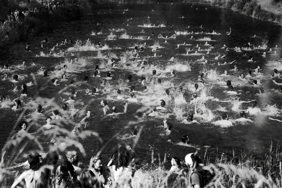 Vue en plongée d’une rivière bondée de dizaines de personnes nageant et éclaboussant l’eau, tandis que d’autres spectateurs se tiennent sur les berges. Scène collective, dense et en mouvement.