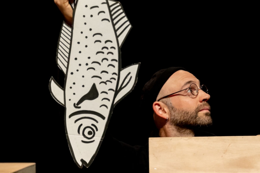 Un homme avec des lunettes se cache, une marionnette en forme de poisson devant lui 