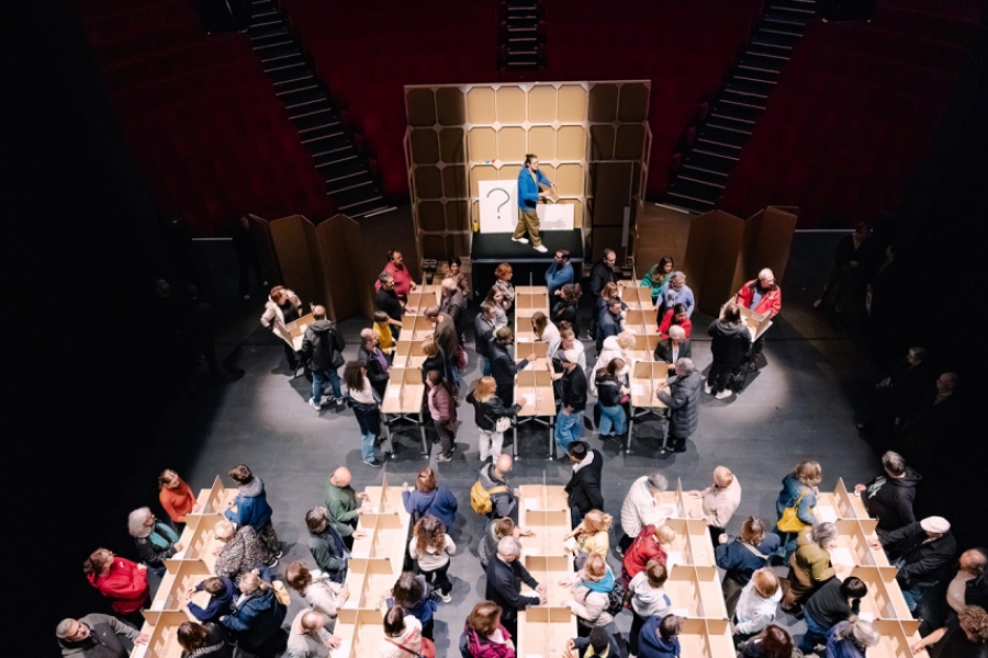 Des personnes sont debout face à face autour de tables, une personne est sur une estrade avec un tableau 