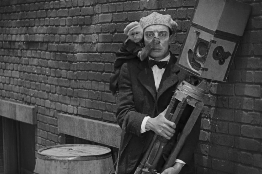 Buster Keaton tient un trépied de caméra, un signe sur ses épaules lui recouvre le nez avec sa main 